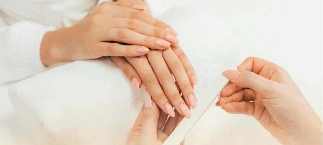 A technician filing a client's nude almond nails over a white towel
